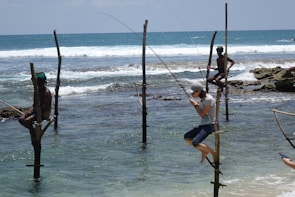 Several individuals are practicing stilt fishing in the ocean, sitting on wooden poles anchored in shallow waters. Waves gently crash in the background, and there is a sense of calm and concentration among the fishermen.