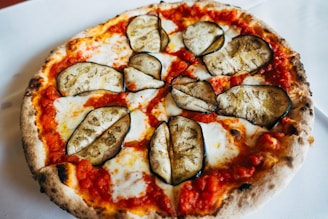 A pizza featuring a thin, crispy crust topped with a layer of rich tomato sauce, melted cheese, and slices of roasted eggplant. The crust has a golden-brown hue with some charred spots, giving it an authentic wood-fired appearance.