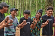A group of five young men are standing in a forest, holding hands and laughing together. They are dressed casually and appear to be enjoying each other's company amidst the tall trees.