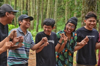 A group of five young men are standing in a forest, holding hands and laughing together. They are dressed casually and appear to be enjoying each other's company amidst the tall trees.