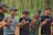 A group of five young men are standing in a forest, holding hands and laughing together. They are dressed casually and appear to be enjoying each other's company amidst the tall trees.