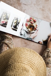 A person holding an open book with images of figs on a plate. The book features close-up photos of figs, including some sliced open to show the interior. The person is wearing a flower-patterned blouse and a woven straw hat. The book is placed on a light marble surface.