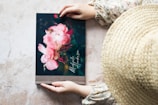 A person wearing a floral-patterned blouse and a straw hat is holding a book with a cover featuring pink flowers and the name 'Michelle Henderson' written on it. The surface beneath the book has a light, textured appearance.