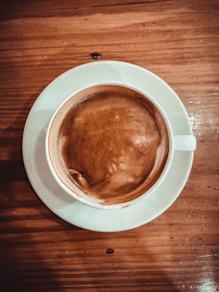 A steaming cup of coffee is placed on a white saucer with a wooden table as the background. The rich, dark liquid has a smooth and creamy texture.