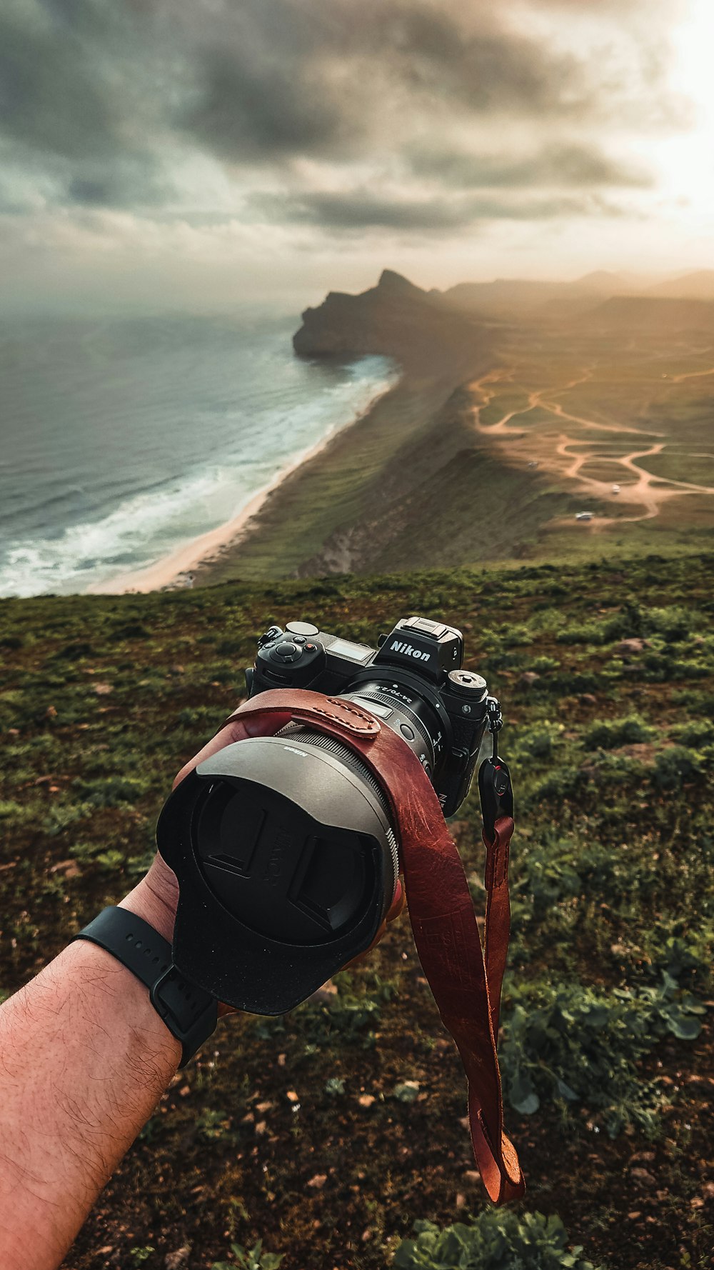 a person's hand holding a camera on a hill overlooking a beach