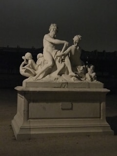 An intricately carved stone sculpture depicting several figures in a classical style, positioned on a pedestal. The dim lighting casts soft shadows, enhancing the contours of the sculpture.