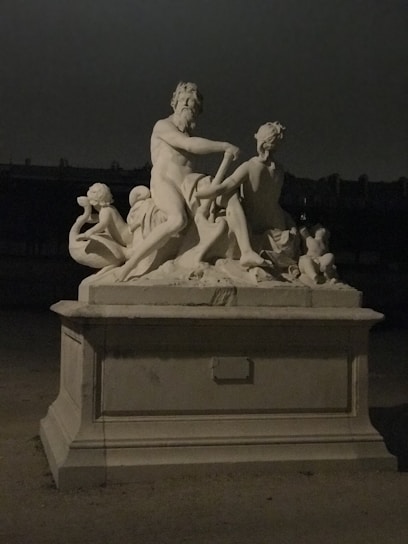 An intricately carved stone sculpture depicting several figures in a classical style, positioned on a pedestal. The dim lighting casts soft shadows, enhancing the contours of the sculpture.