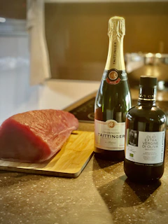 A raw piece of meat is placed on a wooden cutting board next to a bottle of champagne and a bottle of extra virgin olive oil. The scene is set on a kitchen countertop with a stove in the background.