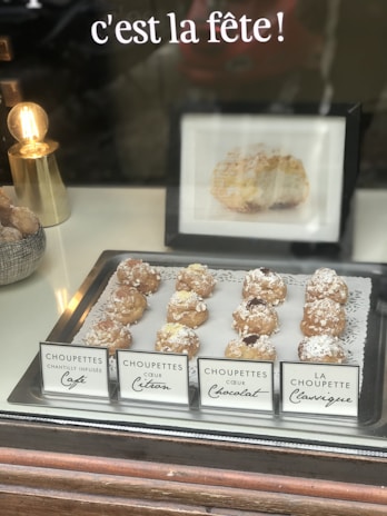 A display of various choux pastry desserts, each labeled with different flavors including café, citron, and chocolat, is set on a silver tray lined with a decorative doily. A small gold lamp with an exposed bulb is positioned to the side, adding a warm glow. In the background, a French phrase 'c'est la fête!' is visible above the pastries.