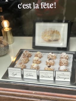 A display of various choux pastry desserts, each labeled with different flavors including café, citron, and chocolat, is set on a silver tray lined with a decorative doily. A small gold lamp with an exposed bulb is positioned to the side, adding a warm glow. In the background, a French phrase 'c'est la fête!' is visible above the pastries.