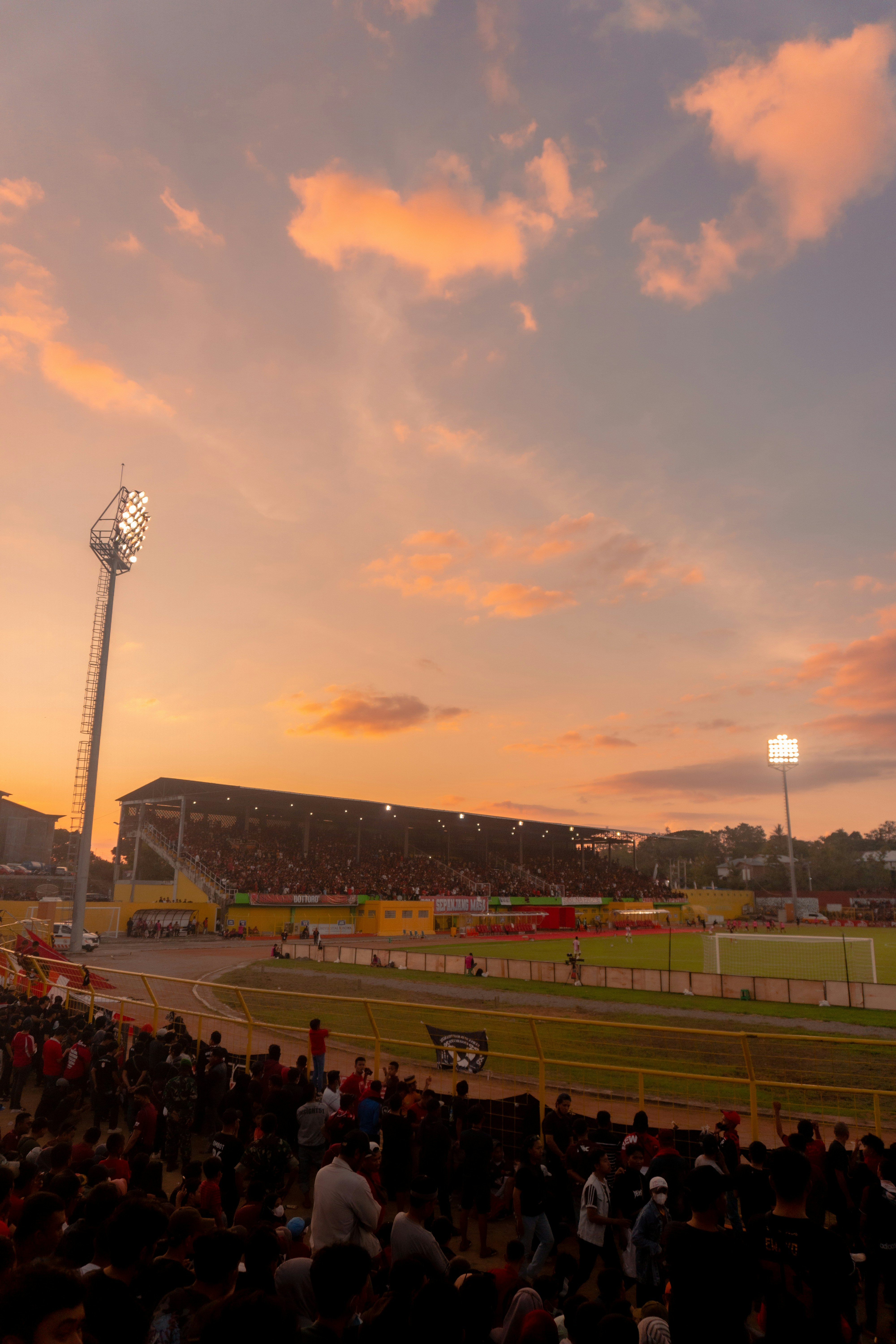 Massive packed outdoor stadium at golden hour, dramatic sky, huge crowd, atmospheric light, deep rich colors