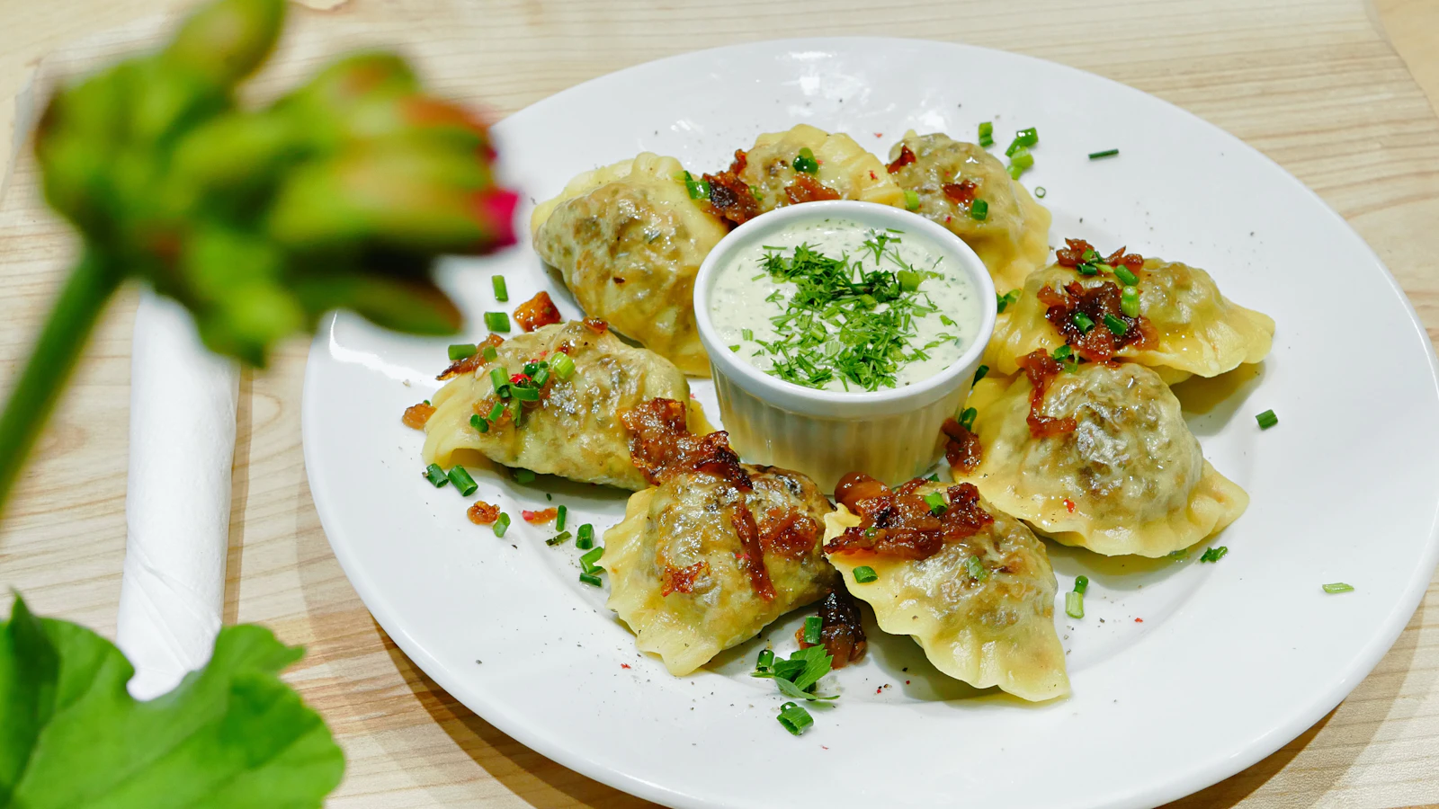 A white plate of golden pan-finished pierogi topped with crispy bacon bits and chives, arranged around a small bowl of herbed sour cream dip