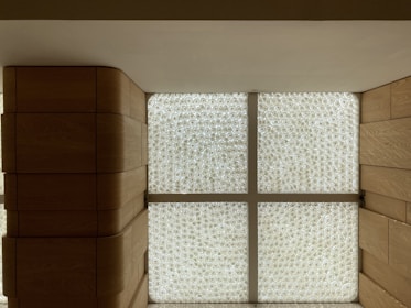 A modern interior with wooden walls and a large, backlit textured glass window panel divided into four sections by cross-shaped framework. The glass features intricate circular patterns, and the wood has a smooth, warm finish.