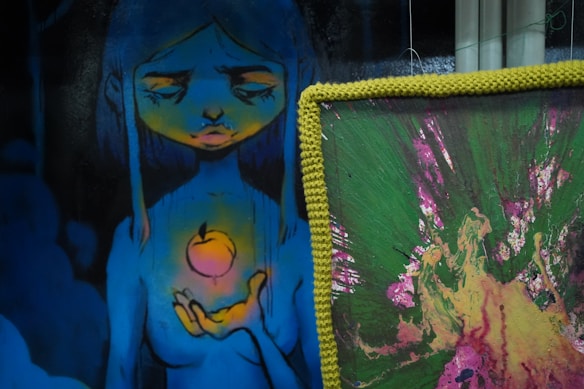 A piece of artwork depicts a blue-toned figure with long hair, holding a glowing peach-colored orb in its hand. The figure has closed eyes and an expression that suggests a contemplative or serene mood. To the right, there is a separate canvas with an abstract design featuring vibrant splashes of green, pink, and yellow, bordered by a yellow knitted frame.