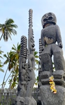 Tall, intricate wooden carvings resembling traditional totem poles are surrounded by palm trees under a partly cloudy sky. The carvings are detailed with expressive faces and patterns. A garland of colorful flowers is draped over one of the statues.
