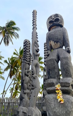 Tall, intricate wooden carvings resembling traditional totem poles are surrounded by palm trees under a partly cloudy sky. The carvings are detailed with expressive faces and patterns. A garland of colorful flowers is draped over one of the statues.