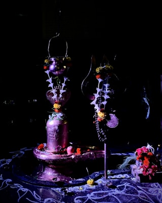 A dimly lit scene features a ceremonial Hindu setup, including a Shiva Lingam adorned with flowers and other ritualistic items. Traditional ornaments, marigold flowers, and leaves surround the central object on a decorated platform. Soft white designs are drawn on the floor, adding to the spiritual ambiance.
