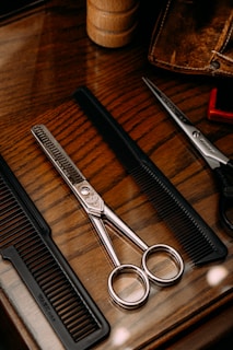 A wooden surface holds a pair of metallic scissors and two black combs. The scissors feature sleek, shiny handles and a textured blade, placed between the combs. A brown leather case and a wooden cylindrical object are also partially visible, adding a rustic aesthetic.