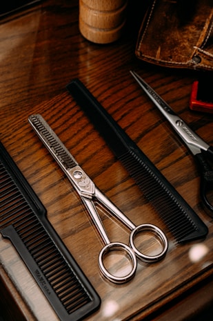 A wooden surface holds a pair of metallic scissors and two black combs. The scissors feature sleek, shiny handles and a textured blade, placed between the combs. A brown leather case and a wooden cylindrical object are also partially visible, adding a rustic aesthetic.