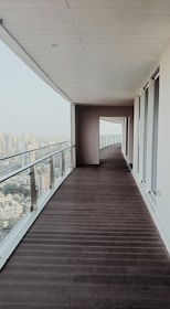 An outdoor balcony with wooden flooring stretches alongside a tall building, offering a panoramic view of a city skyline. The balcony is enclosed with a glass railing and is lined with floor-to-ceiling windows on one side. The scene conveys a modern architectural design and a sense of openness.