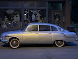 A vintage silver car is parked on the street in front of a small retail store, which has a sign reading 'MOUTON ROYAL.' The store's window displays various clothing items and accessories. The scene is dimly lit, suggesting it is evening or early morning.