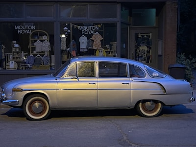 A vintage silver car is parked on the street in front of a small retail store, which has a sign reading 'MOUTON ROYAL.' The store's window displays various clothing items and accessories. The scene is dimly lit, suggesting it is evening or early morning.