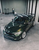 A sleek, dark green sports car is parked in a modern, spacious garage with a polished concrete floor. The car has a sporty design with a prominent rear spoiler and visible branding on the side. The surrounding area includes several storage items such as pallet jacks and a fire extinguisher on the wall.