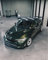 A sleek, dark green sports car is parked in a modern, spacious garage with a polished concrete floor. The car has a sporty design with a prominent rear spoiler and visible branding on the side. The surrounding area includes several storage items such as pallet jacks and a fire extinguisher on the wall.
