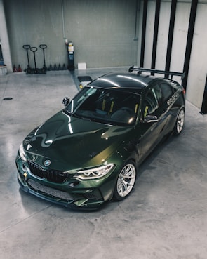 A sleek, dark green sports car is parked in a modern, spacious garage with a polished concrete floor. The car has a sporty design with a prominent rear spoiler and visible branding on the side. The surrounding area includes several storage items such as pallet jacks and a fire extinguisher on the wall.
