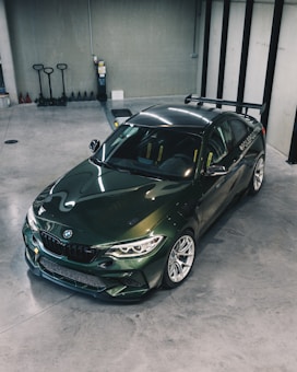 A sleek, dark green sports car is parked in a modern, spacious garage with a polished concrete floor. The car has a sporty design with a prominent rear spoiler and visible branding on the side. The surrounding area includes several storage items such as pallet jacks and a fire extinguisher on the wall.