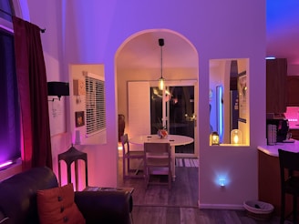 A cozy, dimly lit room with warm and colorful lighting. There is a leather couch with red pillows in the foreground and a small, round dining table with simple wooden chairs in the background. Overhead, a pendant light hangs above the table. To the right, there's a kitchen area with a countertop and a coffee maker. Soft ambient light in shades of purple, pink, and blue adds a relaxing atmosphere. Decorative elements include a vase, a small lamp, and framed artwork on the walls.