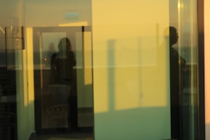 A reflective view through glass panels showing overlapping images of a room's interior and exterior. Shadows of people and architectural elements create a layered and abstract appearance. The soft lighting and blurred reflections contribute to a tranquil, dreamlike atmosphere.