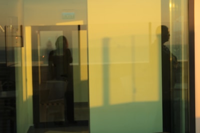 A reflective view through glass panels showing overlapping images of a room's interior and exterior. Shadows of people and architectural elements create a layered and abstract appearance. The soft lighting and blurred reflections contribute to a tranquil, dreamlike atmosphere.