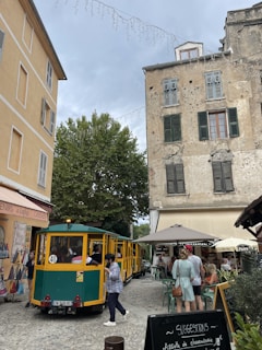 A narrow cobblestone street is lined with old buildings, one of which displays a worn facade with closed shutters. A bright yellow and green tourist train is parked along the street with several people boarding and walking around. Outdoor cafes with umbrellas and seated patrons add to the lively atmosphere of the quaint setting.