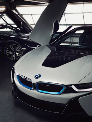 A sleek, modern car with a distinctive design and upward-opening doors is parked indoors. The car features a glossy white exterior with blue accents around the grille and emblems. Another vehicle is partially visible in the background, highlighting the luxurious setting.