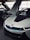 A sleek, modern car with a distinctive design and upward-opening doors is parked indoors. The car features a glossy white exterior with blue accents around the grille and emblems. Another vehicle is partially visible in the background, highlighting the luxurious setting.