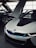 A sleek, modern car with a distinctive design and upward-opening doors is parked indoors. The car features a glossy white exterior with blue accents around the grille and emblems. Another vehicle is partially visible in the background, highlighting the luxurious setting.