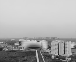 Aerial view of a modern urban landscape featuring a large, circular, multi-story building and several rectangular buildings nearby. The area appears to be an office or residential complex surrounded by open land and distant high-rise buildings. The image is in black and white, giving it a stark and contemporary feel.