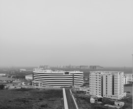 Aerial view of a modern urban landscape featuring a large, circular, multi-story building and several rectangular buildings nearby. The area appears to be an office or residential complex surrounded by open land and distant high-rise buildings. The image is in black and white, giving it a stark and contemporary feel.
