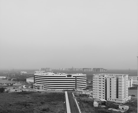 Aerial view of a modern urban landscape featuring a large, circular, multi-story building and several rectangular buildings nearby. The area appears to be an office or residential complex surrounded by open land and distant high-rise buildings. The image is in black and white, giving it a stark and contemporary feel.