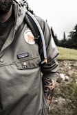 A person wearing a green jacket with a patch featuring a bear and the word 'Patagonia' is visible. A tattoo is partially visible on the individual's right forearm. A small panda pin is attached to the jacket pocket. The person has a hydration pack with a tube resting on the shoulder. The background consists of a grassy area and a forest with tall trees under an overcast sky.