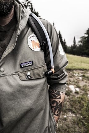 A person wearing a green jacket with a patch featuring a bear and the word 'Patagonia' is visible. A tattoo is partially visible on the individual's right forearm. A small panda pin is attached to the jacket pocket. The person has a hydration pack with a tube resting on the shoulder. The background consists of a grassy area and a forest with tall trees under an overcast sky.