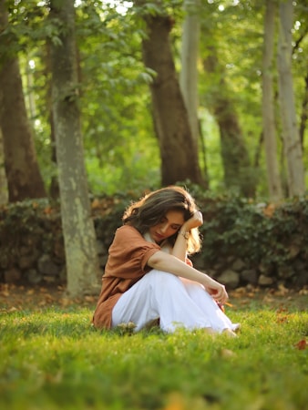 A young woman sits on the grass in a serene forest setting, surrounded by lush trees. She has a thoughtful expression, resting her head on her hand, wearing a brown shirt and white pants. The light filters softly through the leaves, creating a warm, peaceful ambiance.