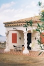 A quaint Mediterranean-style house with a tiled roof and a balcony overlooking a body of water. The house features twisted columns, red shutters, and a patterned patio. A table and chairs are placed outside, suggesting a cozy outdoor seating area. Lush greenery partially frames the scene, adding to the serene atmosphere.