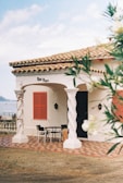 A quaint Mediterranean-style house with a tiled roof and a balcony overlooking a body of water. The house features twisted columns, red shutters, and a patterned patio. A table and chairs are placed outside, suggesting a cozy outdoor seating area. Lush greenery partially frames the scene, adding to the serene atmosphere.