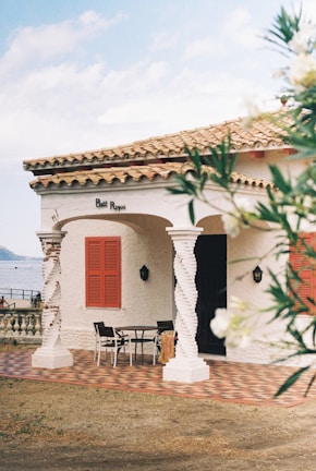 A quaint Mediterranean-style house with a tiled roof and a balcony overlooking a body of water. The house features twisted columns, red shutters, and a patterned patio. A table and chairs are placed outside, suggesting a cozy outdoor seating area. Lush greenery partially frames the scene, adding to the serene atmosphere.