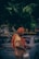 A man dressed in traditional attire with an orange turban and robe stands near a parked car under the shade of a large tree. He holds a plastic bag in one hand. The background includes a yellow bank sign and other urban elements.