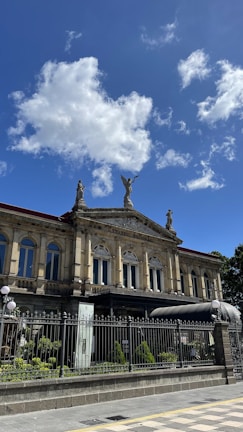 A historic theater building with ornate architectural details, statues on the rooftop, and an iron fence in front. The structure is set against a bright blue sky with fluffy clouds.