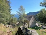 A rustic stone house is nestled in a lush, mountainous landscape. The structure is surrounded by tall trees and large rocks, with a clear blue sky overhead. The scene conveys a sense of tranquility and isolation, with the mountains visible in the background.