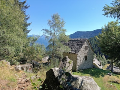 A rustic stone house is nestled in a lush, mountainous landscape. The structure is surrounded by tall trees and large rocks, with a clear blue sky overhead. The scene conveys a sense of tranquility and isolation, with the mountains visible in the background.